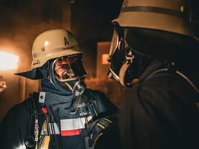 Zwei Feuerwehrmänner unter Atemschutz stehen sich gegenüber. Im Hintergrund ist eine Rauchentwicklung erkennbar.
