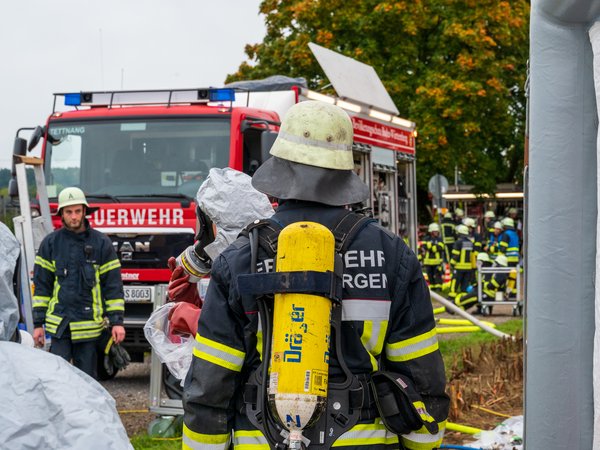 Ein Feuerwehrmann der Feuerwehr Langnnargen steht mit Atemschutz am Dekonplatz und wartet darauf, dekontaminiert zu werden. Um ihn herum sind mehrere Kräfte in Schutzanzügen und Masken.